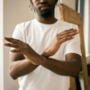 Crop unrecognizable bearded African American male in white t shirt demonstrating no gesture with hands while standing in light room